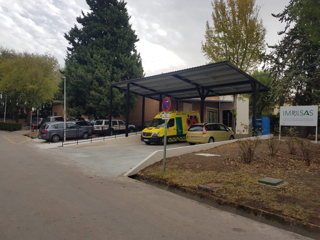 Un aparcamiento cubierto con una ambulancia amarilla y un coche delante de un edificio. Al fondo se ven árboles y un cartel que dice "IMRUSAS".